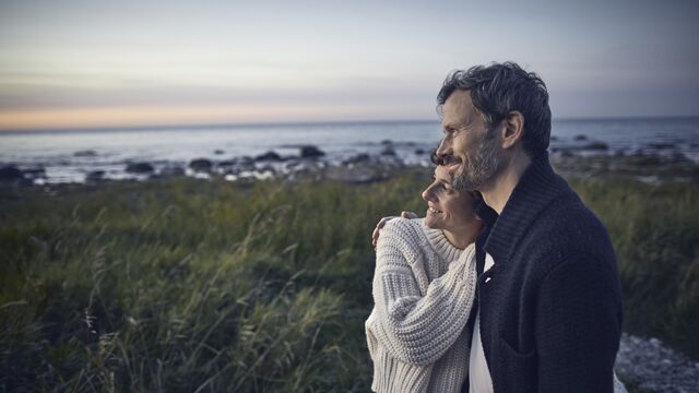 Pariskunta seisoo rannalla toisiinsa nojaten ja katsoo auringonlaskua.