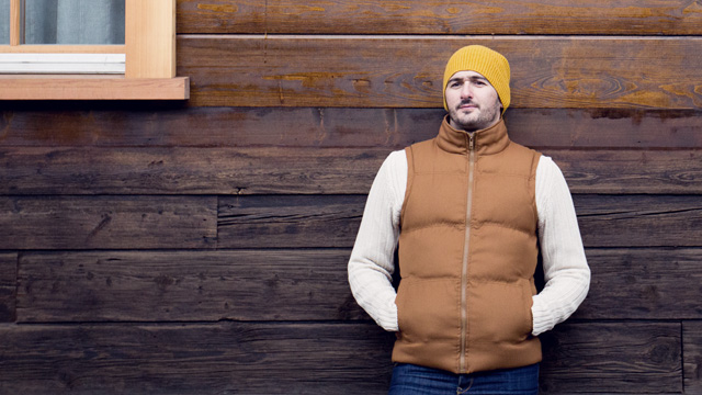 Man with brown jacket leaning against a wooden house 