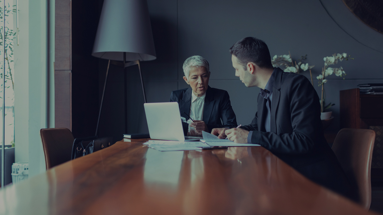 Two people in a meeting in front of a computer and a pile with paper