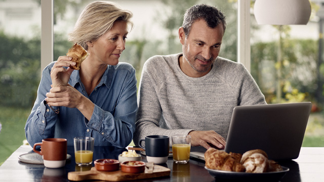 Par hjemme som smiler og spiser foran laptop.